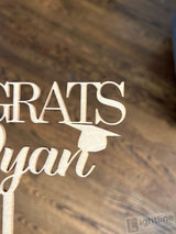 Wooden graduation-themed decorative sign with 'GRATS' and a graduation cap on a wooden surface.
